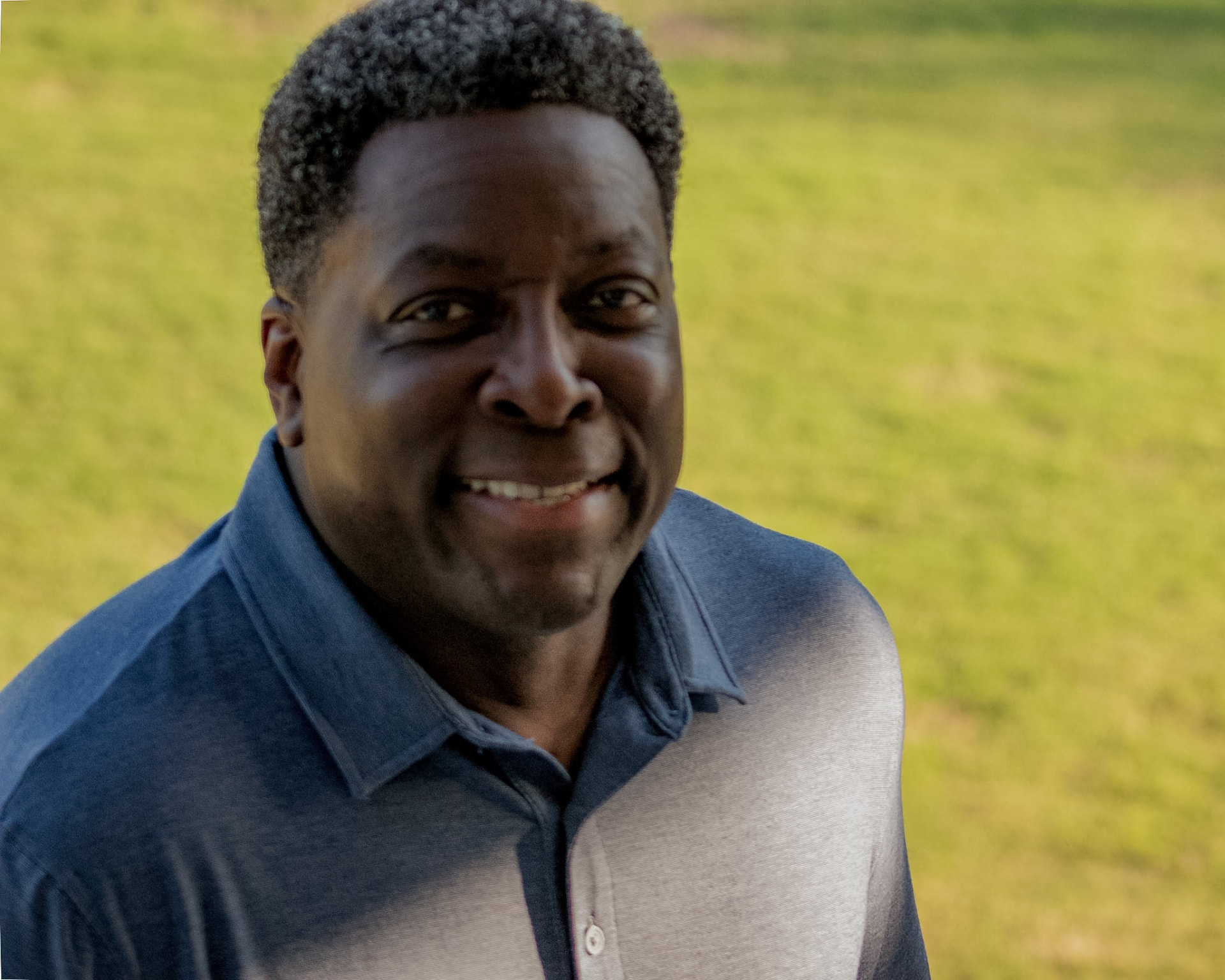Close portrait of Dave London smiling, with a sunlit green field behind him.
