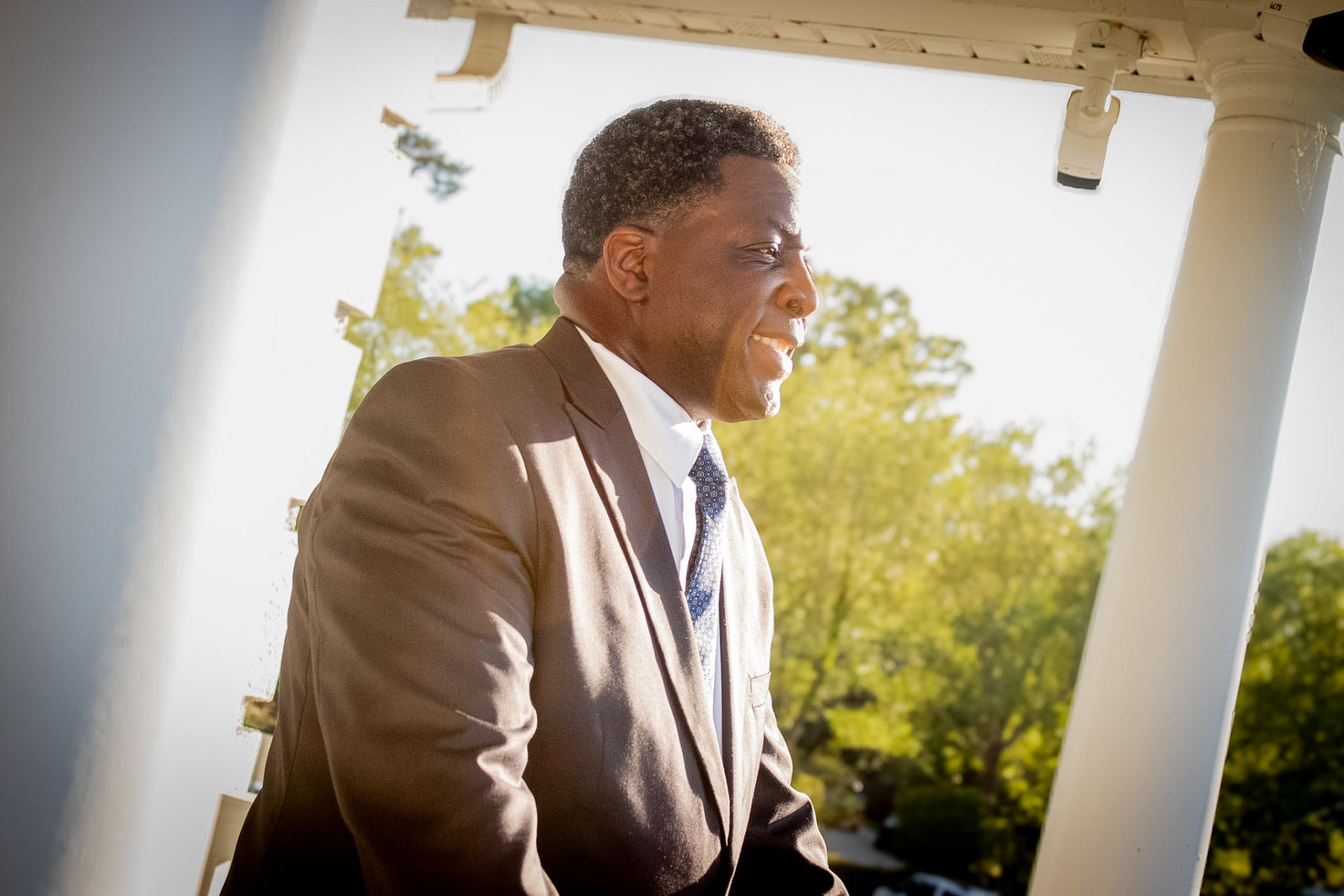 Dave London in a dark suit and tie, backlit by afternoon sun on a columned porch, looking off to his right.